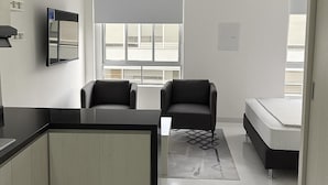 A modern, compact room with a kitchenette, a bed, two chairs, and a large window providing natural light.