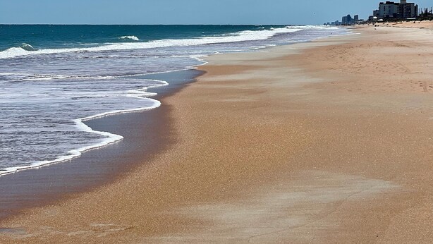 Ein weiter Sandstrand mit sanften Wellen, einem klaren blauen Himmel und Gebäuden in der Ferne.