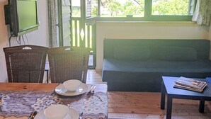 Una habitación con un sofá verde, una mesa de comedor con mantel floreado, una silla de mimbre, un televisor en la pared y una ventana con vistas a zonas verdes y edificios.
