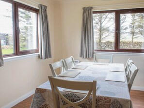 Comedor con mesa dispuesta para seis comensales, ventanas grandes con cortinas y vista a árboles y un muro de piedra.