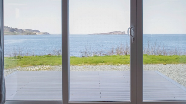 Blick durch eine Glastür auf eine Terrasse mit Blick auf ein ruhiges Meer und entfernte Küste.