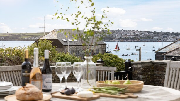 A table set with food and drinks, overlooking a harbor with boats and a town in the background.