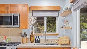A kitchen with wooden cabinets, a stainless steel microwave, a sink, and a window with a view of greenery. - NEW! Volcano Tea House Cottage 2-HotTub-FirePlace (Volcano)