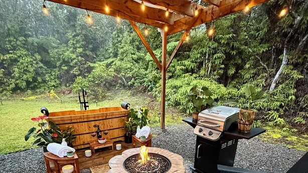 An outdoor patio with a fire pit, wooden furniture, and a grill, surrounded by a natural landscape. - NEW! Volcano Tea House Cottage 2-HotTub-FirePlace (Volcano)