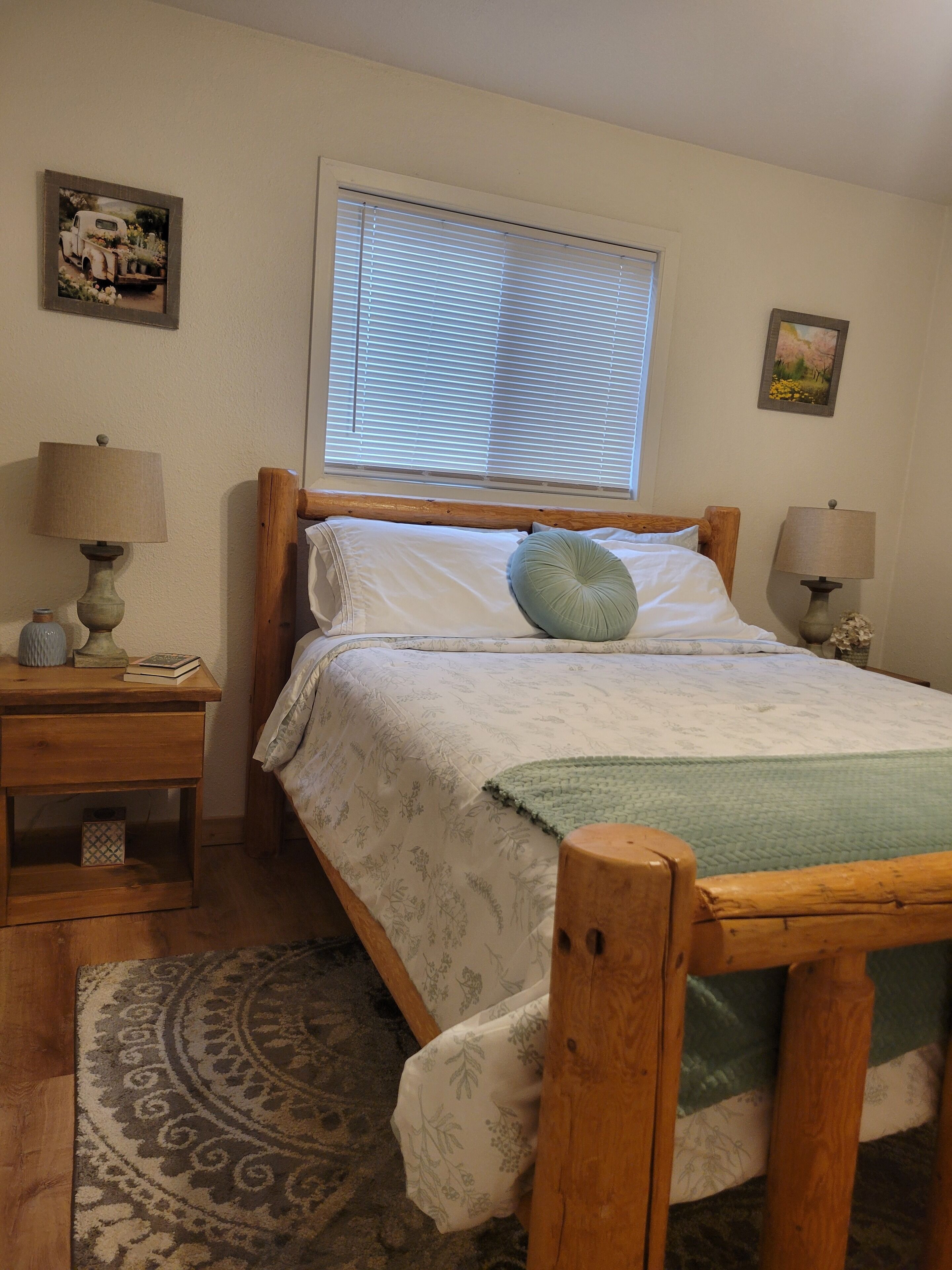 A bedroom with a wooden bed, two bedside tables with lamps, a window with blinds, and a patterned rug.