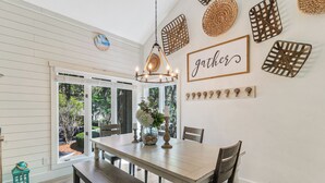 Ein Essbereich mit Holztisch, Bänken und einer Chandelüster. Dekorative Wandbehänge und Blick auf Grünflächen.