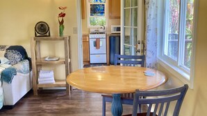A dining area with a wooden table and blue chairs, a kitchen in the background, and a view of greenery outside.