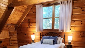 A cozy wooden cabin bedroom with a bed, two bedside tables with lamps, a bench, and a floral rug.