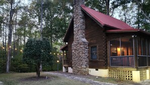 A rustic cabin with a stone chimney, wooden exterior, and a porch with a view of the surrounding forest.