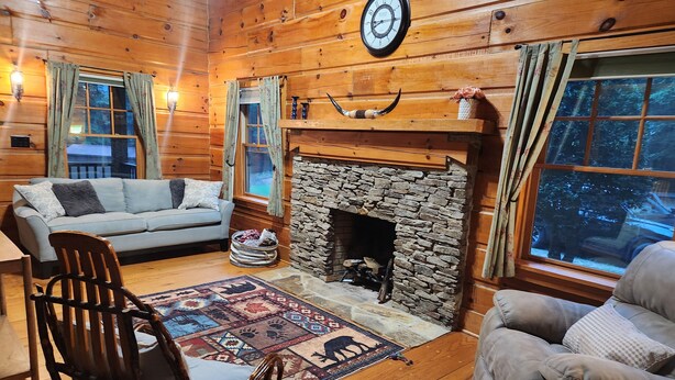 A cozy cabin interior with a stone fireplace, wooden walls, a sofa, a chair, and a rug.