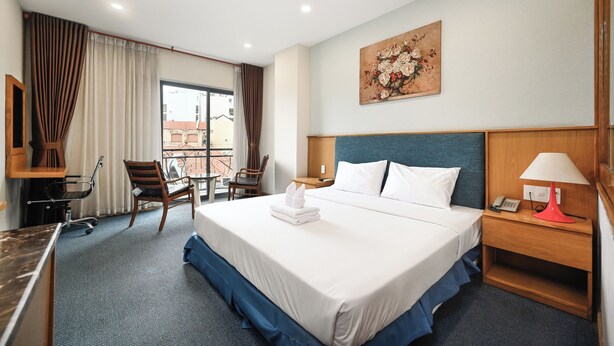 A hotel room with a large bed, a desk with a chair, a small table, a telephone, and a painting on the wall. - Sunshine Airport Hotel (Ho Chi Minh City)