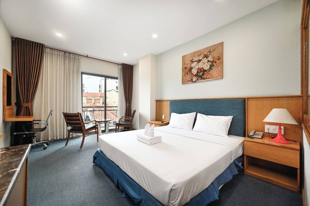 Habitación de hotel con una cama grande, un escritorio con silla, una mesita, un teléfono y un cuadro en la pared.