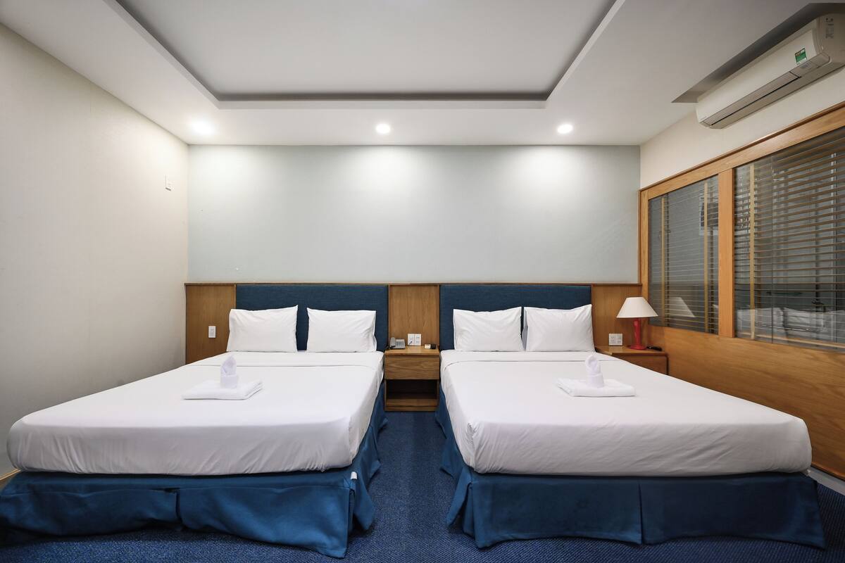 Habitación de hotel con dos camas, cabecera de madera, mesita de noche con lámpara y ventana con persianas.