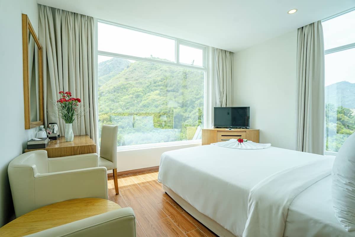 Habitació d'hotel amb un llit, una televisió, una taula de treball amb una cadira, una finestra amb cortines i vistes a una muntanya.