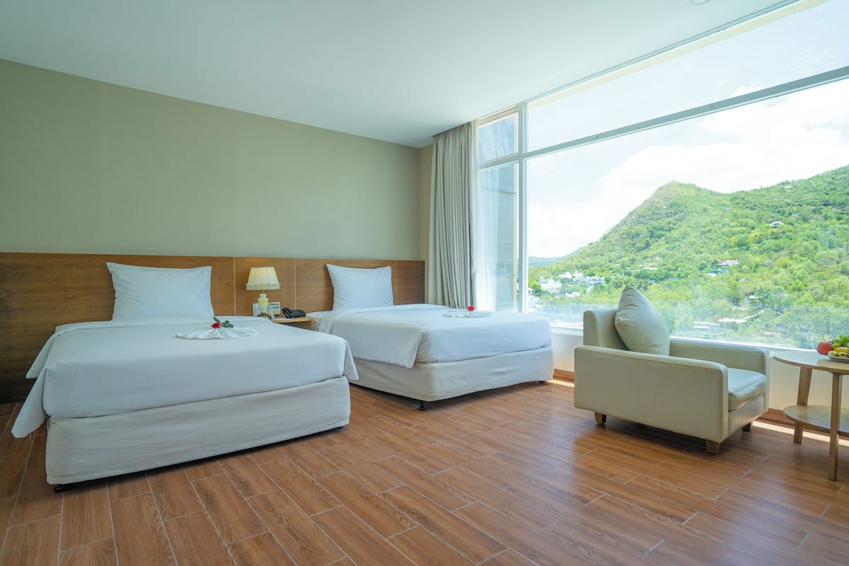 Habitació d'hotel amb dos llits, una cadira, una taula petita i vista a una muntanya verda.