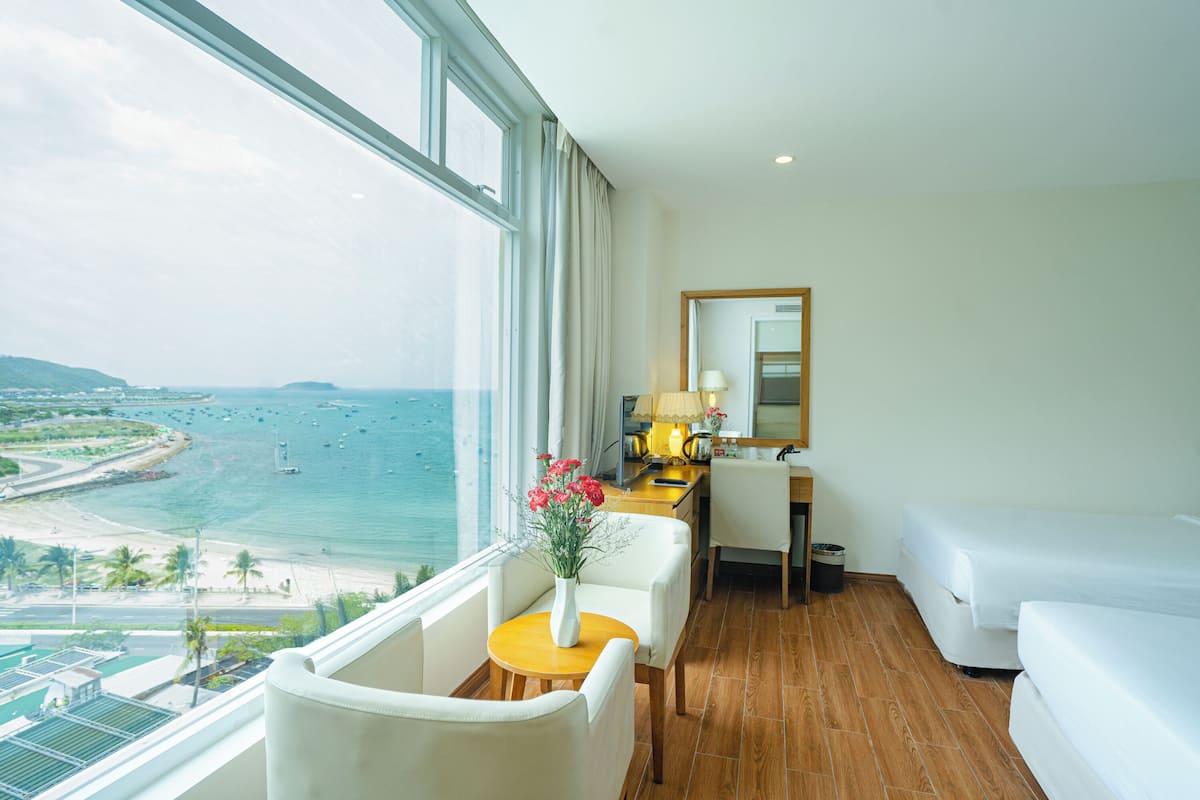 Habitació d'hotel amb una finestra gran que ofereix vistes a la platja i a l'oceà. Hi ha una taula rodona petita amb un ram de flors, dues butaques blanques, un paviment de fusta i un sofà blanc.