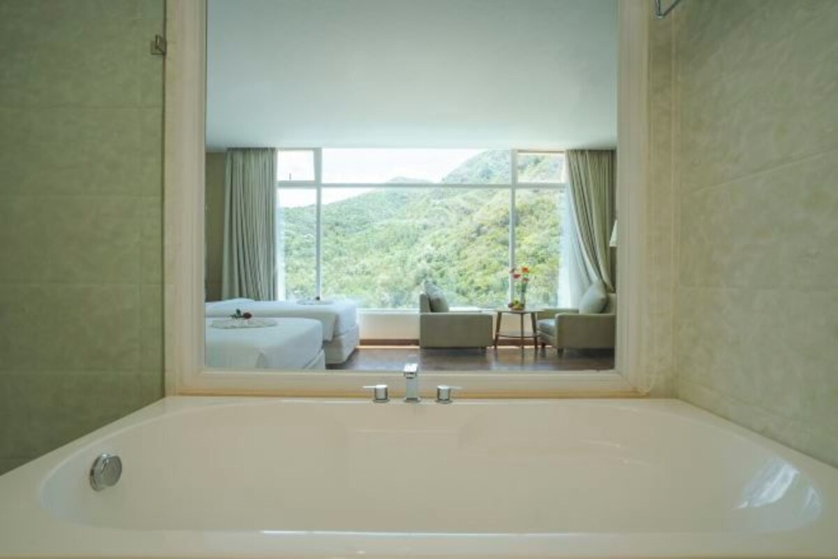 Un baño moderno con una bañera grande, vista a un dormitorio con cama y una mesita, y cortinas en la ventana.