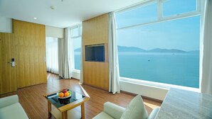 Habitación de hotel moderna con un amplio ventanal que ofrece vistas al mar, un televisor de pantalla plana en la pared y una pequeña mesa con frutas y un control remoto.