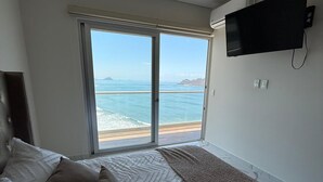 Quarto com cama, televisão fixada na parede e grande janela com vista para o mar.