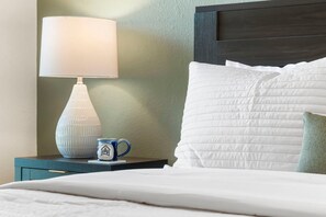 A neatly made bed with white bedding, a white lamp on a bedside table, and a small blue mug with a logo.