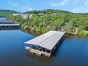 A floating structure with a covered deck, surrounded by calm water, with buildings and trees in the background.
