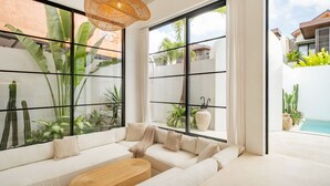 A modern living room with a large window, a woven light fixture, and a view of a pool and greenery. - Casa Blanche Canggu (Canggu)