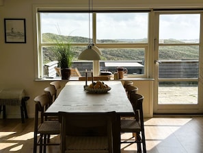 A dining area with a long wooden table, chairs, a potted plant, and a view of the outdoors through large windows.
