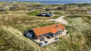 A modern house with a red roof, surrounded by grassy dunes, with a view of the sea in the background.