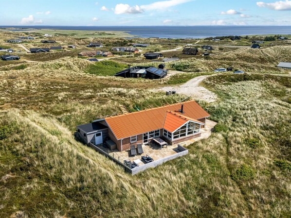 Een modern huis met een rood dak, omgeven door grasduinen, met uitzicht op zee.