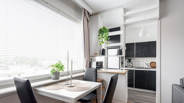 Cocina moderna con armarios blancos, un microondas y un lavamanos. Hay un comedor con mesa y sillas, y una planta en el alféizar de la ventana.