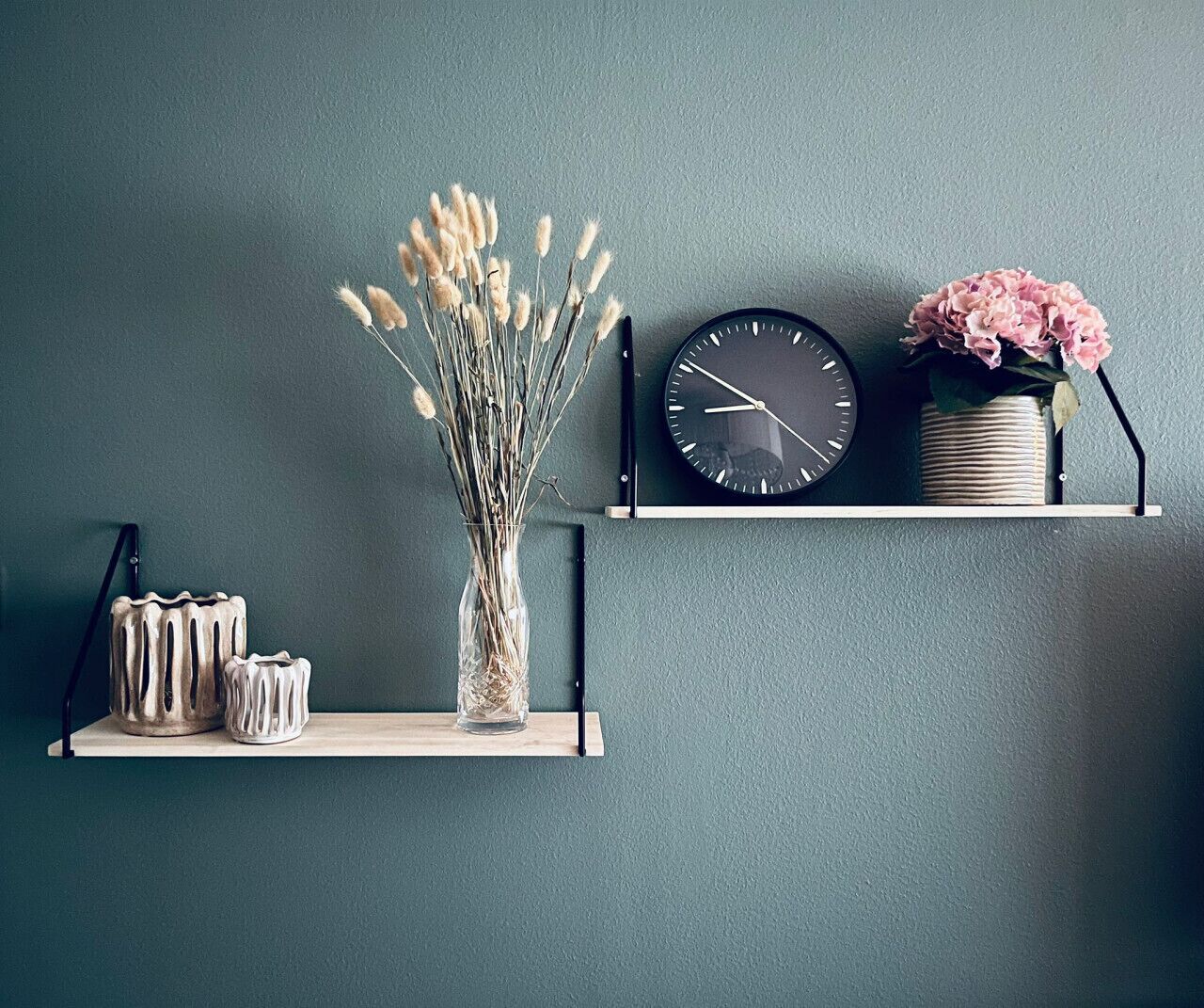 Un reloj de pared, un florero con flores secas y dos recipientes de cerámica sobre una estantería frente a una pared color verde azulado.