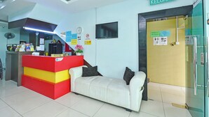 A reception area with a red and yellow desk, a white sofa, a clock, and a television on the wall. - De Mawardah Inn Hotel (Malacca City)