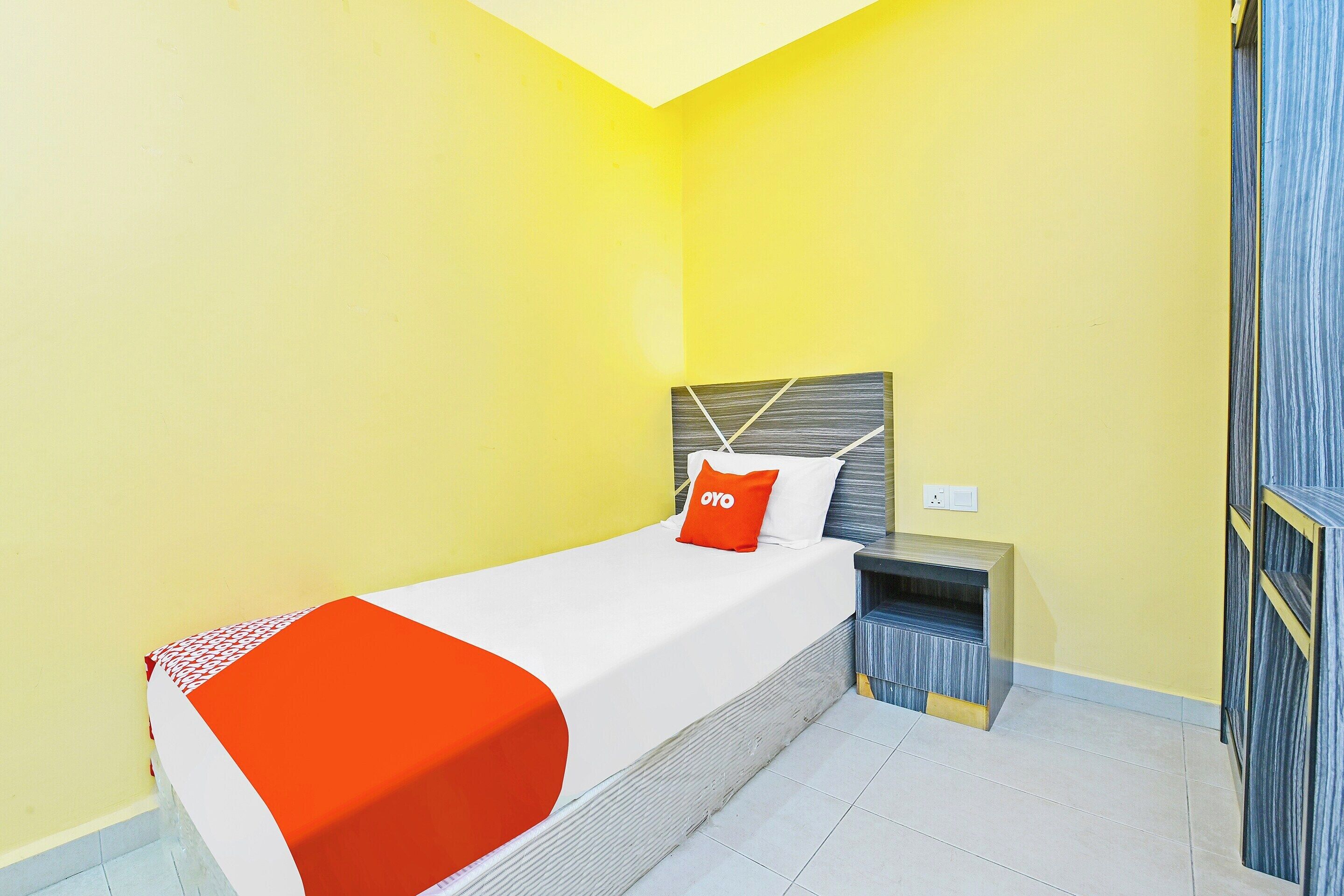 A single bed with a white and orange bedspread, a red pillow with "OYO" text, a wooden headboard, and a nightstand with a lamp.