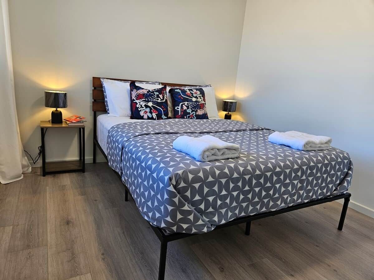 A single bed with a geometric patterned bedspread, two pillows with a floral design, two bedside lamps, and a small wooden nightstand with a magazine.