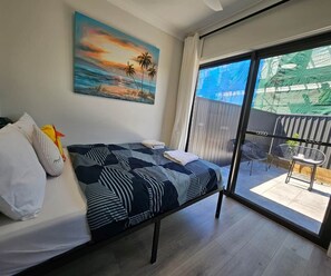 Een slaapkamer met een bed, kussens, een schilderij en een schuifdeur die leidt naar een buitenruimte met een tafel en stoelen.