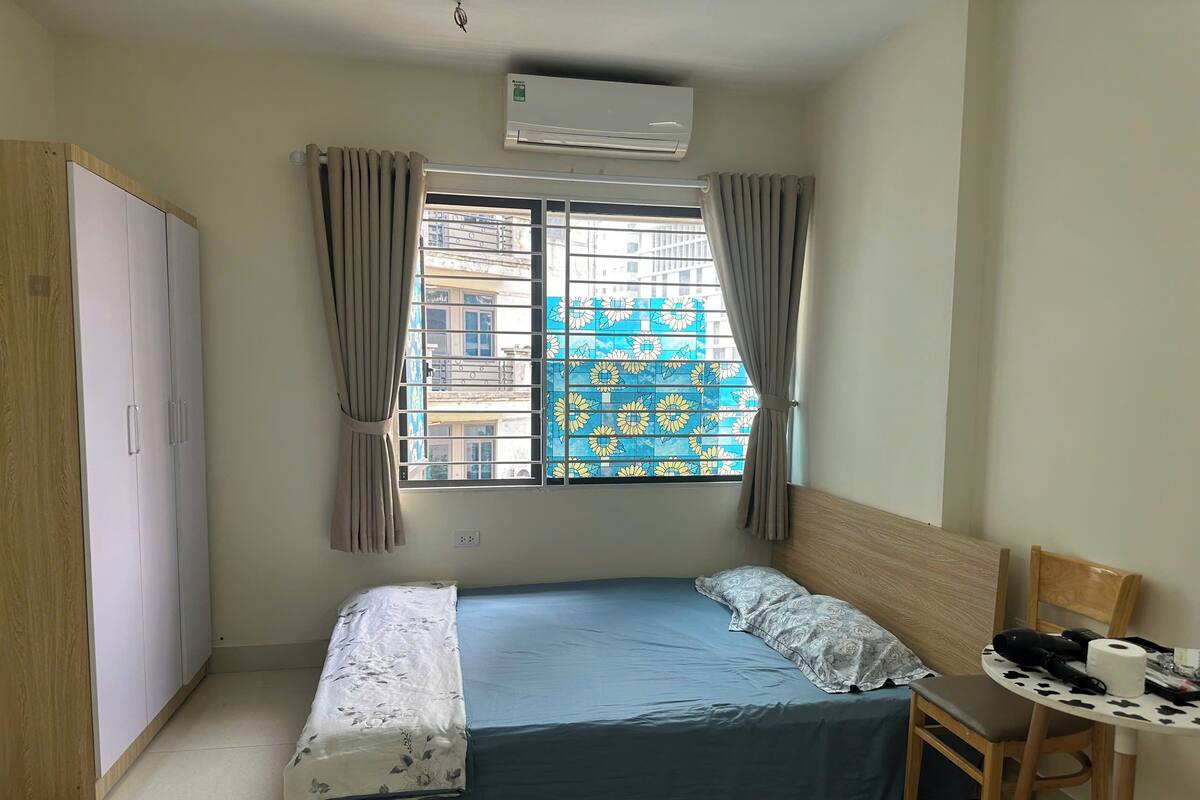 Une chambre à coucher comprenant un lit, une chaise, une table, un climatiseur et une fenêtre avec des rideaux.