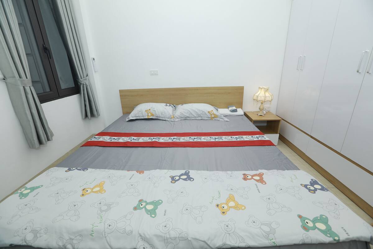 Une chambre à coucher avec un lit, des oreillers, une table de chevet avec une lampe, une armoire et une fenêtre avec des rideaux.