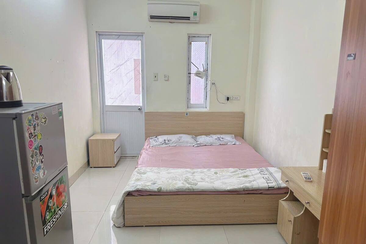 Une chambre à coucher avec un lit, un bureau en bois, un réfrigérateur et deux fenêtres.
