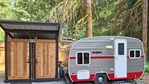 A vintage RV parked next to a wooden shed with a metal roof. - LITTLE BETTY WiFi, A/C,FirePit, BBQ (Ashford)