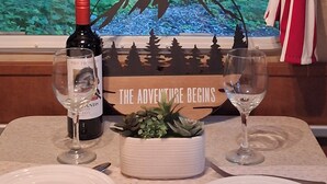 A dining table with two wine glasses, a bottle of wine, and a decorative sign with a forest scene. - LITTLE BETTY WiFi, A/C,FirePit, BBQ (Ashford)