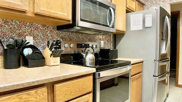 A kitchen with wooden cabinets, a stainless steel microwave, a kettle, and a refrigerator.