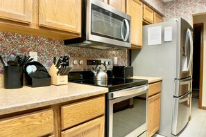 A kitchen with wooden cabinets, a stainless steel microwave, a kettle, and a refrigerator.