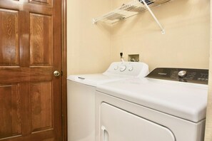 Un área de lavandería con una lavadora y secadora Whirlpool blancas, una puerta de madera y un estante metálico sobre la secadora.