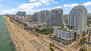A coastal cityscape with high-rise buildings, a beach with sun loungers, and a clear sky. - Gorgeous Jr Suite on Pool and Lanai Level of Hotel (Fort Lauderdale)