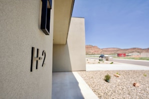 L’extérieur d’un bâtiment, avec une plaque indiquant les numéros « 1-2 », une structure en béton et une zone recouverte de gravier avec une végétation clairsemée.