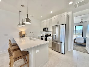A modern kitchen with white cabinetry, stainless steel appliances, pendant lighting, and a view to the outside.