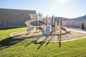 Un parc infantil amb tobogans i estructures per escalar, envoltat d'una zona verda i un edifici al fons.