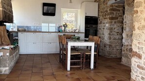 A rustic kitchen with stone walls, a tiled floor, a wooden dining table with chairs, a built-in oven, and a hanging light fixture. - Waterfront house with large walled garden (Île-aux-Moines)