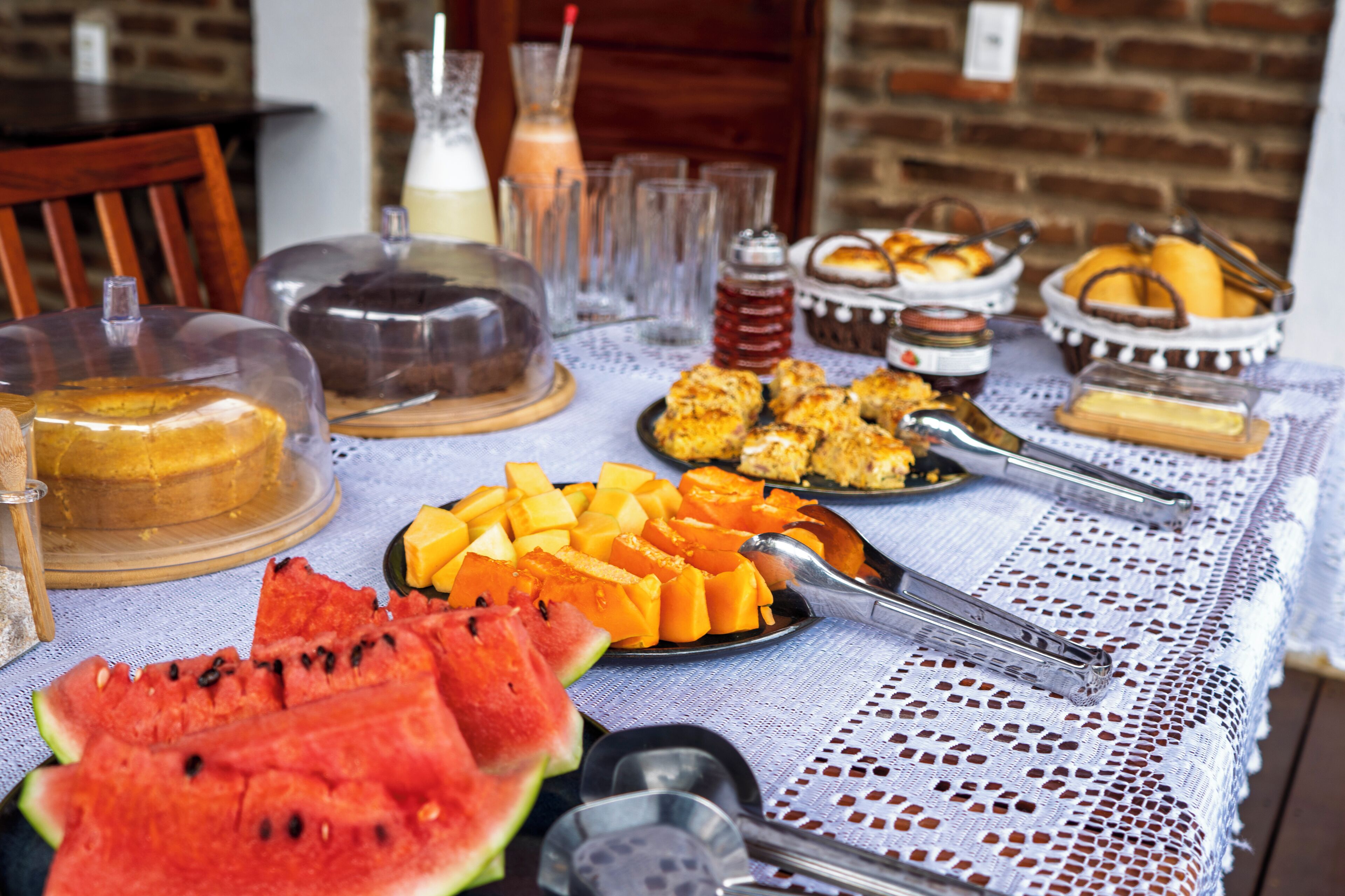 Uma mesa com diversos alimentos, incluindo melancia, queijo e pão, coberta por uma toalha de renda.