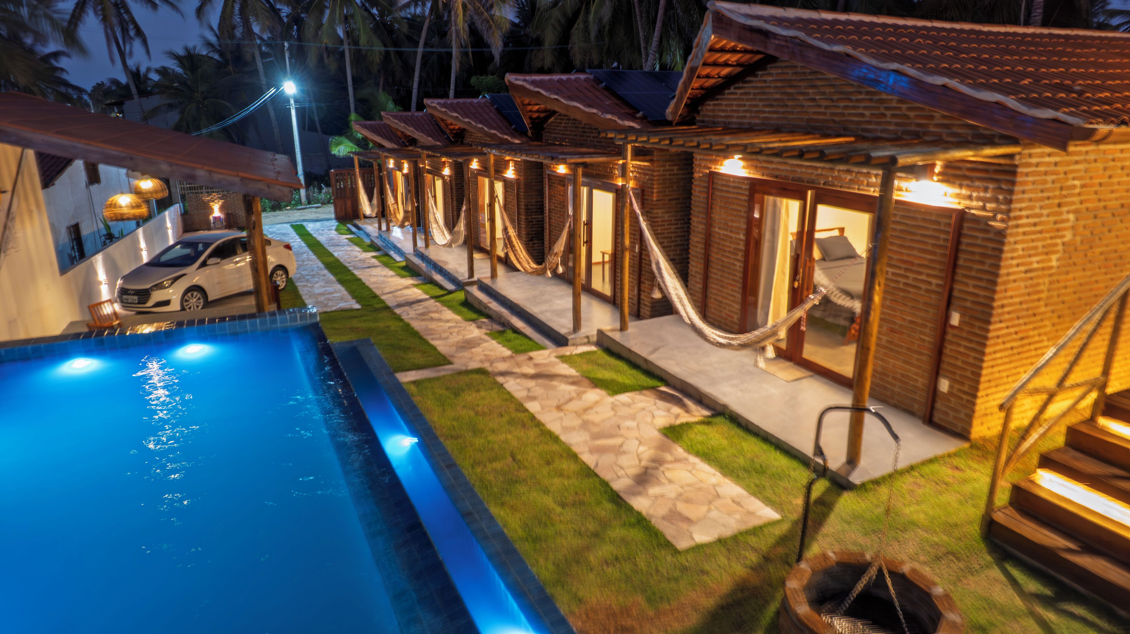 A poolside area with a swimming pool, a car, and a row of brick buildings with hammocks.